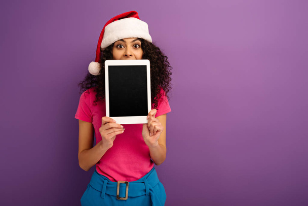 веселая девушка смешанной расы в шляпе Санты показывает цифровой планшет с пустым экраном на фиолетовом фоне
 - Фото, изображение