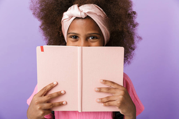 Optimistic young african girl covering face behind the book - Foto, Imagen