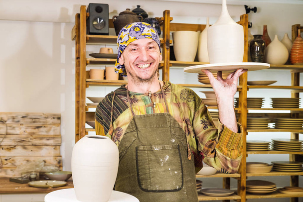 glimlachende mannelijke pottenbakker meester met aardewerk kunstproducten in zijn werkplaats - Foto, afbeelding