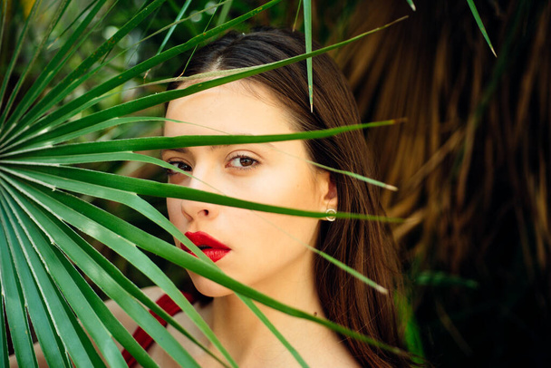 Beautiful woman holding a leaf of a palm tree. Facial treatment. Tanned skin. Skin care. Brunette beautiful fashion model girl face. Woman with palm trees. Beauty red lips make up photo. - Photo, Image