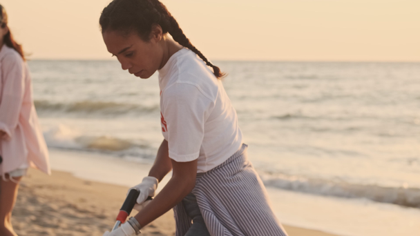 Ελκυστικοί πολυεθνικοί εθελοντές που φορούν λευκά μπλουζάκια καθαρίζουν την παραλία από πλαστικά με σακούλες σκουπιδιών στην παραλία την αυγή - Πλάνα, βίντεο
