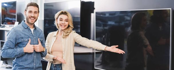 panoramisch schot van vriendje tonen houdt van en glimlachende vriendin wijzend met de hand naar nieuwe tv in huis apparaat winkel  - Foto, afbeelding