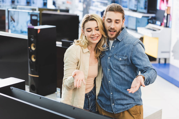 novio y novia sonriente señalando con las manos en la nueva televisión en la tienda de electrodomésticos
  - Foto, Imagen
