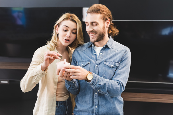 smiling boyfriend holding piggy bank and girlfriend putting coin into it in home appliance store  - Photo, Image