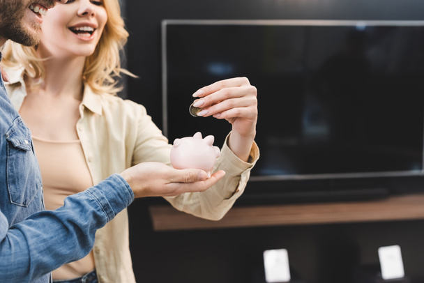 cropped view of smiling boyfriend holding piggy bank and girlfriend putting coin into it in home appliance store  - Fotografie, Obrázek