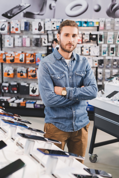 przystojny mężczyzna z skrzyżowanymi ramionami stojący w pobliżu nowych smartfonów w sklepie AGD  - Zdjęcie, obraz