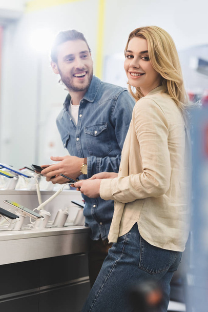smiling boyfriend and girlfriend holding new smartphones in home appliance store  - Foto, Bild