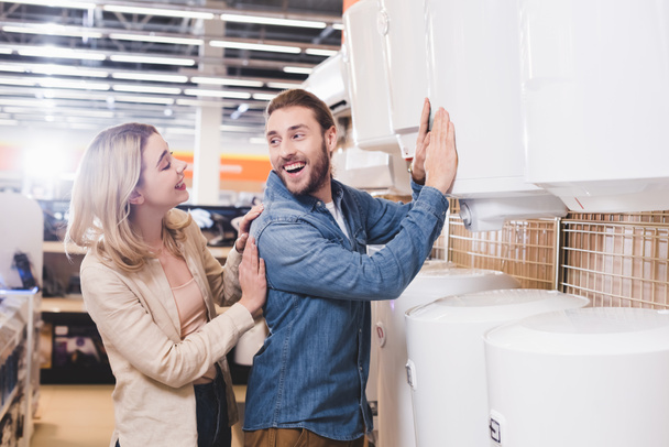 smiling boyfriend touching boiler and looking at girlfriend in home appliance store  - Photo, Image