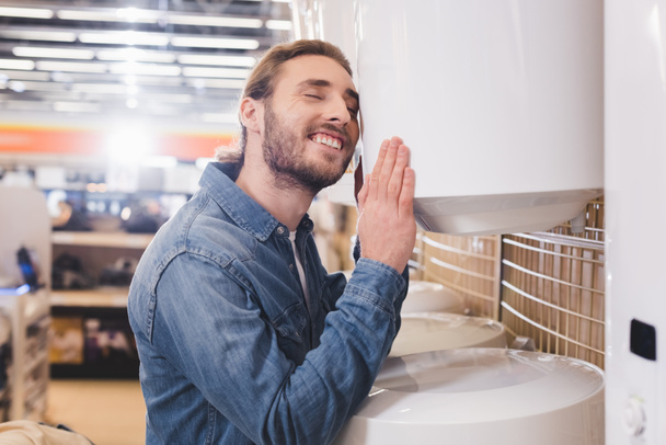 smiling man with closed eyes touching boiler in home appliance store  - Photo, Image