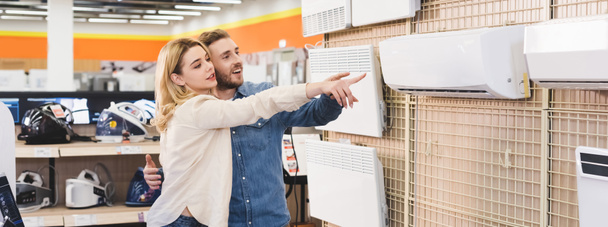 panoramic shot of smiling boyfriend and girlfriend pointing with fingers at air conditioner in home appliance store  - Foto, immagini