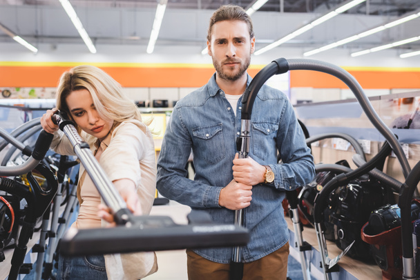 serious boyfriend and girlfriend holding vacuum cleaners in home appliance store  - 写真・画像