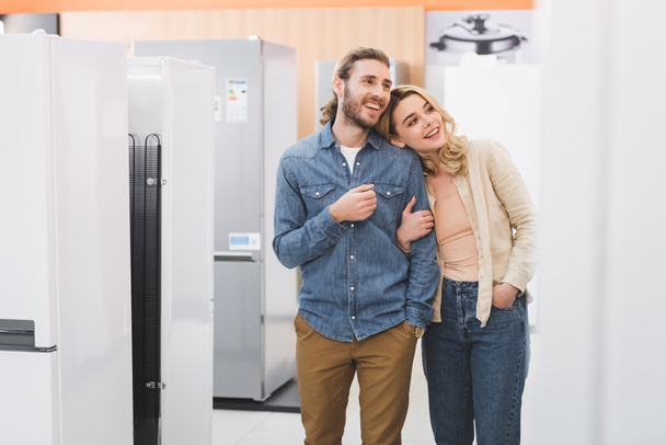 smiling boyfriend and girlfriend looking at fridge in home appliance store  - Foto, Imagen
