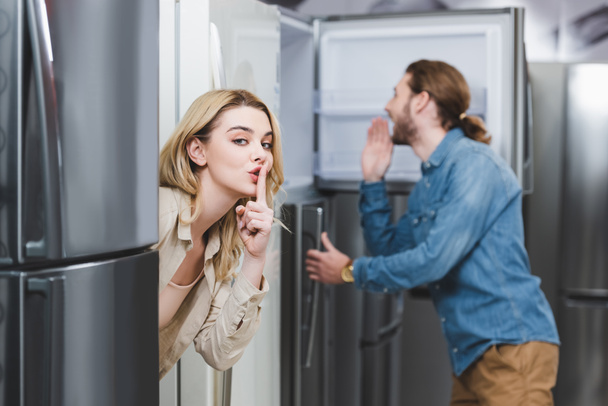 selective focus of girlfriend showing shh and boyfriend looking at fridge on background in home appliance store  - Photo, Image
