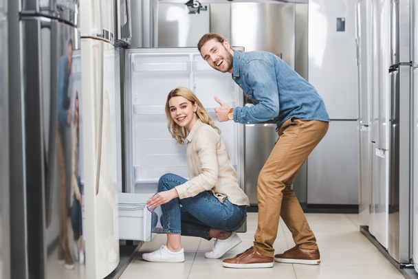 smiling boyfriend showing like and girlfriend sitting near new fridge in home appliance store  - 写真・画像