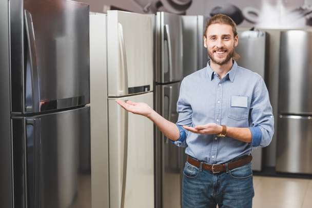 Lächelnder Berater zeigt mit den Händen auf Kühlschrank im Haushaltsgerätehaus  - Foto, Bild
