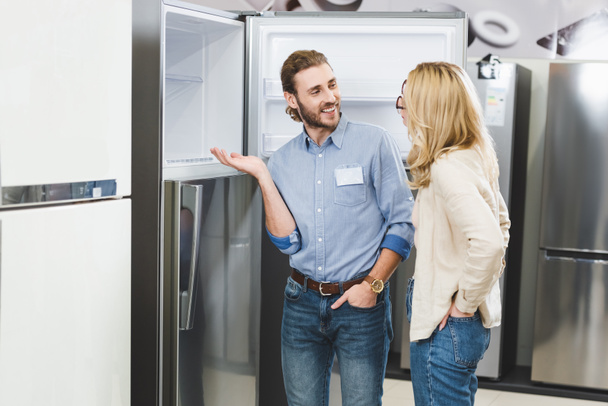 smiling consultant pointing with hand at fridge and talking with woman in home appliance store  - 写真・画像