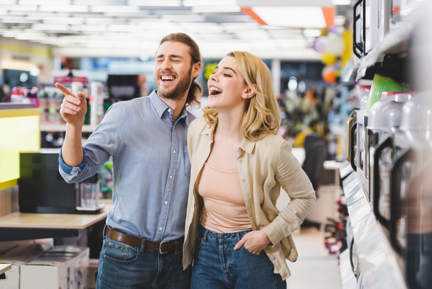 konzultant ukazující prstem a usmívající se žena stojící u mikrovlnky v obchodě s domácími spotřebiči  - Fotografie, Obrázek