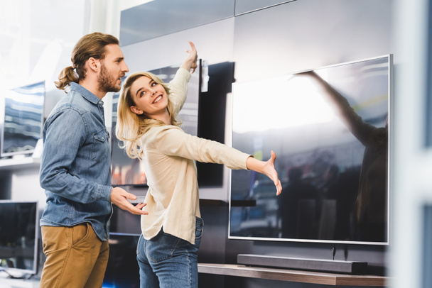 smiling girlfriend pointing with hands at tv and looking at shocked boyfriend in home appliance store  - Fotoğraf, Görsel