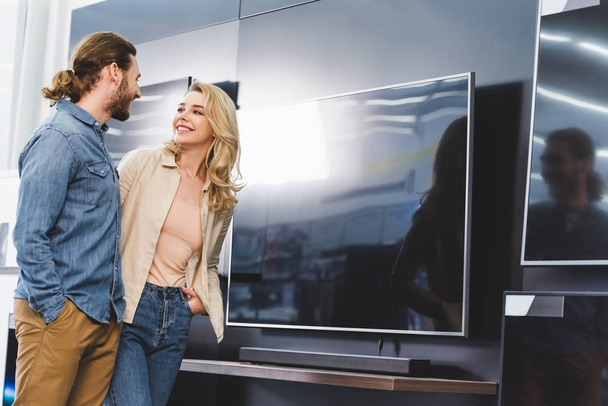 smiling boyfriend and girlfriend standing near tv in home appliance store  - Фото, изображение