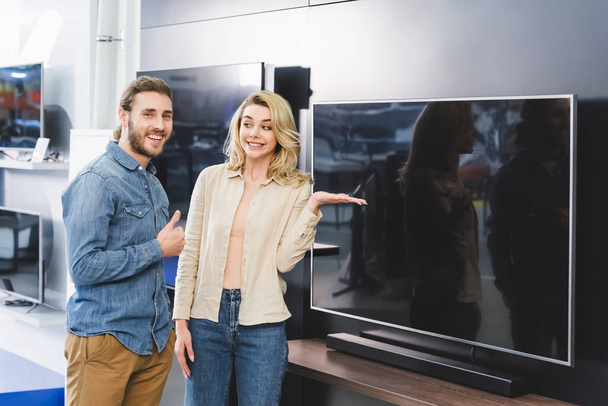 smiling boyfriend showing like and girlfriend pointing with hand at tv in home appliance store  - Zdjęcie, obraz