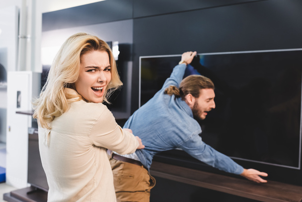 selective focus of angry girlfriend pulling boyfriend with tv in home appliance store  - Photo, Image