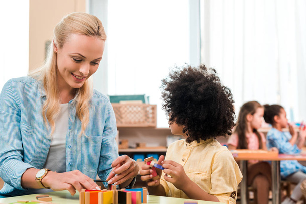 Selektivní zaměření na usmívající se učitel a dítě hrát vzdělávací hru s dětmi v pozadí ve škole Montessori  - Fotografie, Obrázek