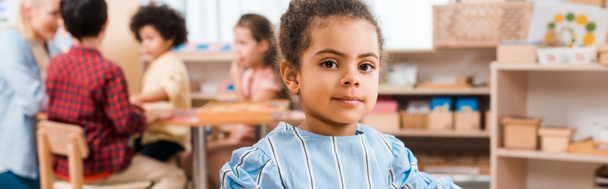 Enfoque selectivo del niño afroamericano mirando a la cámara con el maestro y los niños en el fondo en la escuela montessori
 - Foto, Imagen