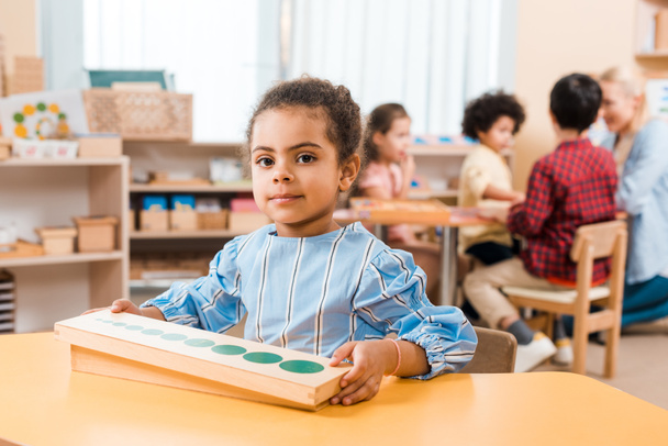 Selectieve focus van kind met houten spel kijken naar camera en leraar met kinderen op de achtergrond in Montessori school - Foto, afbeelding