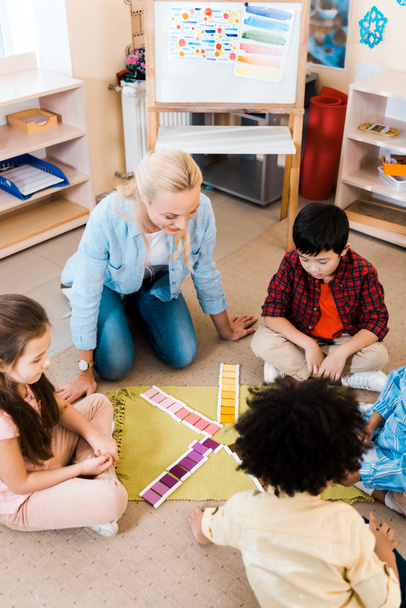 Montessori okulunda yerde oturan çocukların ve renkli oyun katlayan öğretmenlerin genel bakış açısı beyazdır. - Fotoğraf, Görsel