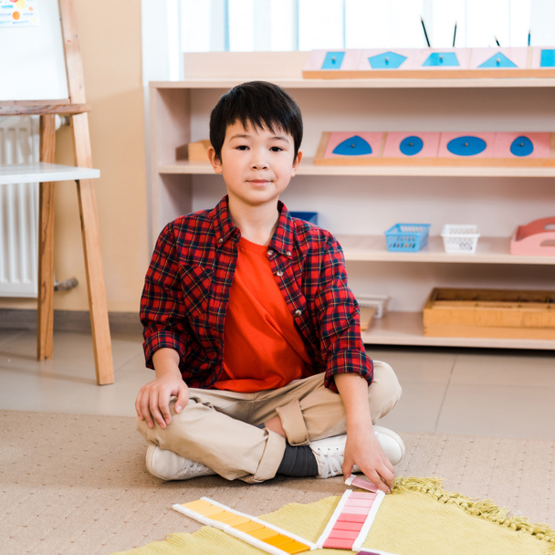 asiatisches Kind blickt in die Kamera, während es buntes Spiel auf dem Boden in der Montessori-Schule faltet - Foto, Bild