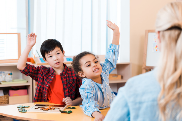 Selective focus of children with raised hands and teacher during lesson in montessori class - 写真・画像