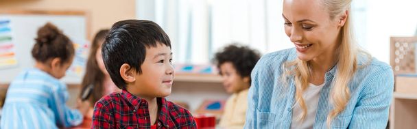 Prise de vue panoramique d'un enseignant souriant et d'un enfant avec des enfants en arrière-plan à l'école montessori
 - Photo, image