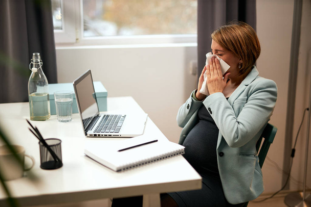 Sick caucasian pregnant businesswoman sitting in her office and blowing her nose. - Foto, Bild