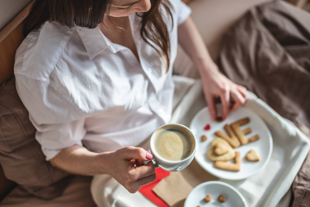 Mladá žena má romantickou snídani v ranní posteli. Káva a slovo láska ze sušenek. Překvapení na Valentýna - Fotografie, Obrázek