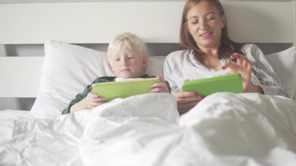Mom and son lie in bed in the morning and check social networks on tablets - Footage, Video