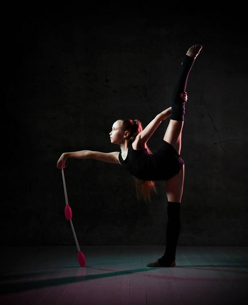 Young smiling girl gymnast in black sport body and uppers standing and holding two pink gymnastic maces in hand - Foto, imagen