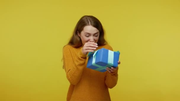 Attractive excited girl with ginger hair in pullover catching present and shaking box with interest, curious about gift, expressing extreme happiness. indoor studio shot isolated on yellow background - Filmati, video