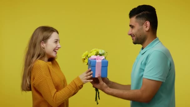 Handsome brunette man giving present and flowers to beautiful ginger girlfriend, she shaking gift with interest, guy gesturing no, i won't say what's in box. studio shot isolated on yellow background - Filmati, video