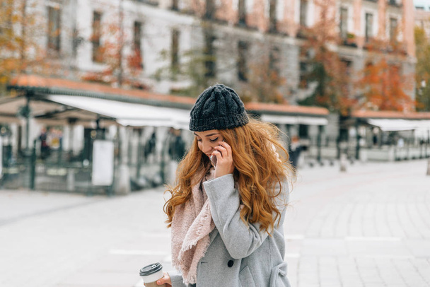 Mujer rubia joven con ropa de invierno habla en el teléfono móvil mientras lleva una taza de café en la otra mano
 - Foto, Imagen