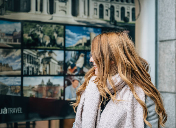 Una rubia de pelo largo con abrigo mira a una ventana de tienda de una agencia de viajes. - Foto, imagen
