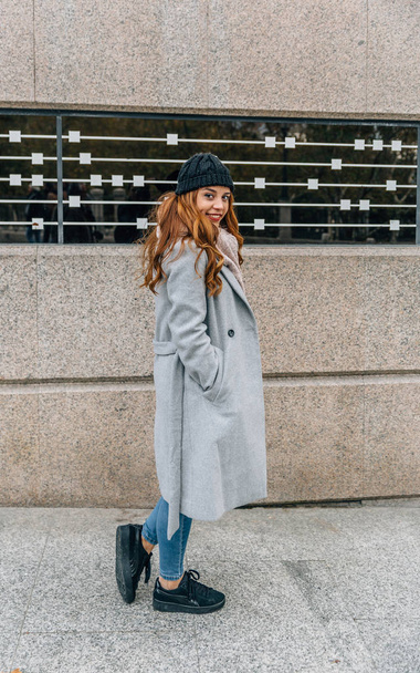 Urban style blonde girl dressed in gray coat and woolen hat walks smiling on a street - Фото, изображение