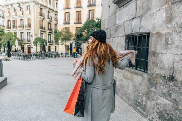 Девочка-блондинка с большим розовым шарфом на спине во время прогулки по городу с несколькими сумками для покупок - Фото, изображение