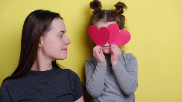 Young mother and cute child daughter holding red hearts sitting on yellow background waving hands look at camera talking at webcam say hello greeting friend or relatives. Blogger concept - Footage, Video