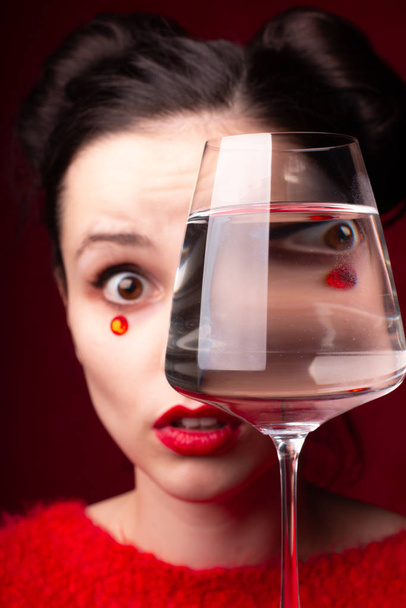 emotional girl in red with red lips and rhinestones on her face holds a glass in her hands - Foto, afbeelding