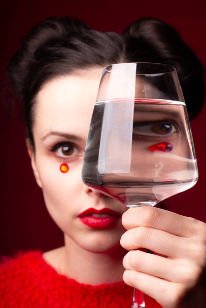 эмоциональная девушка в красном с красными губами и стразами на лице держит стакан в руках
 - Фото, изображение
