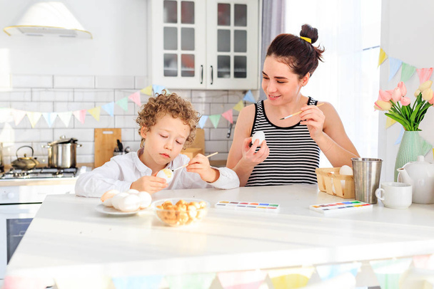 happy young dark-haired mom and her curly blond son paint eggs for easter in the white kitchen in the Scandinavian style - Zdjęcie, obraz