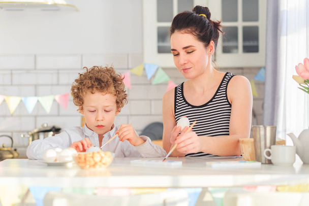 happy young dark-haired mom and her curly blond son paint eggs for easter in the white kitchen in the Scandinavian style - 写真・画像