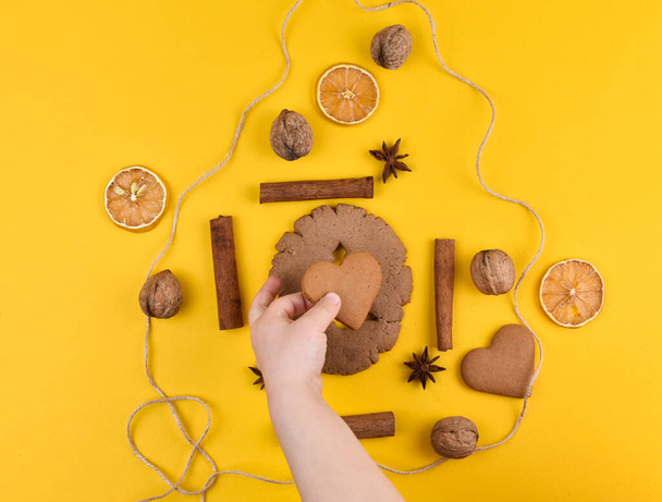 Dětská ručička vloží koláčky ve tvaru srdce do středu kruhu. Perníková sušenka na žlutém pozadí. Vanilka, oříšky, sušený citron, hvězda anýzu. - Fotografie, Obrázek