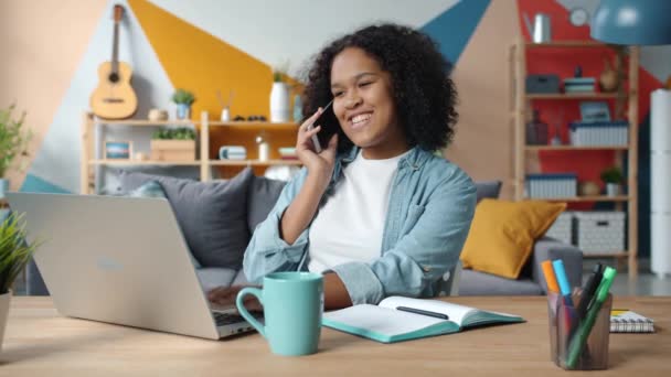 Onnellinen nuori nainen puhuu matkapuhelimella hymyillen työskentelemällä kannettavan tietokoneen kanssa kotona
 - Materiaali, video