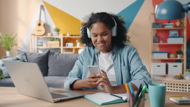 Joyful Afro-American lady touching smartphone screen listening to music in headphones - Кадри, відео